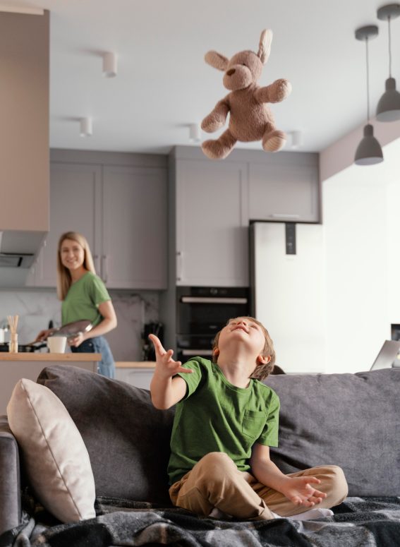 Mutter in offener Wohnkueche und Kind spielt auf Wohnzimmercouch